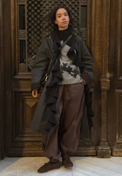 Mujer con abrigo largo oscuro, suéter gris claro con diseño floral negro, pantalones marrones holgados y zapatos marrones, de pie junto a una puerta de madera.