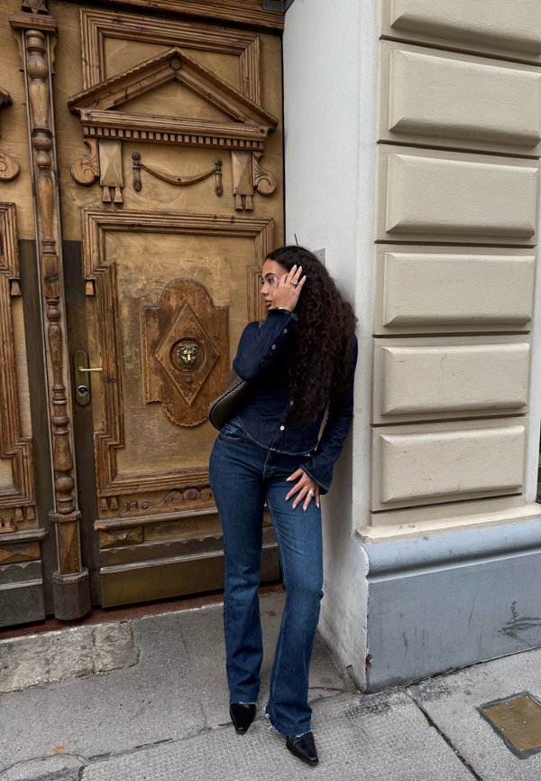 Cămașă din denim și blugi evazați de o nuanță închisă, cu un design ajustat, alături de o ușă din lemn maro texturată, cu detalii ornamentale.