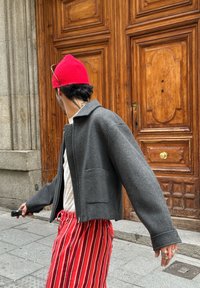 Γκρι κομμένο σακάκι με φερμουάρ μπροστά και μια τσέπη, σε συνδυασμό με κόκκινες και λευκές ριγέ παντελόνες. Ένα φωτεινό κόκκινο σκούφο και δαχτυλίδια είναι ορατά.