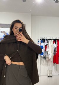 Cardigã canelado castanho usado sobre um top escuro, combinado com calças cinzentas. O fundo apresenta várias roupas em cabides.