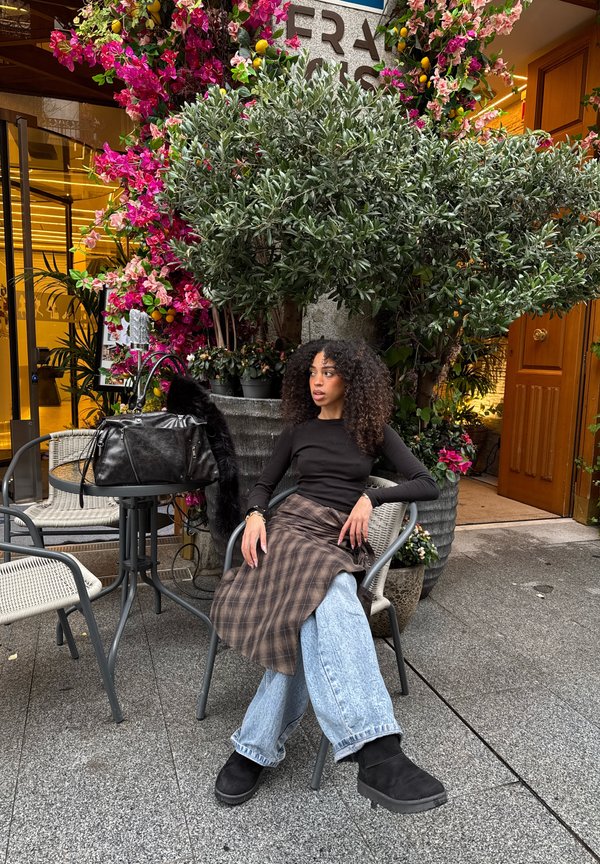 Žena s kudrnatými vlasy sedící na židli venku u zářivě růžových květin a zelených rostlin, oblečená v černém topu, kostkované sukni a širokých džínách.