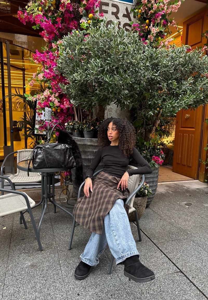 Femme aux cheveux bouclés assise sur une chaise en plein air à côté de fleurs roses vives et de plantes vertes, portant un haut noir, une jupe à carreaux et un large jean.