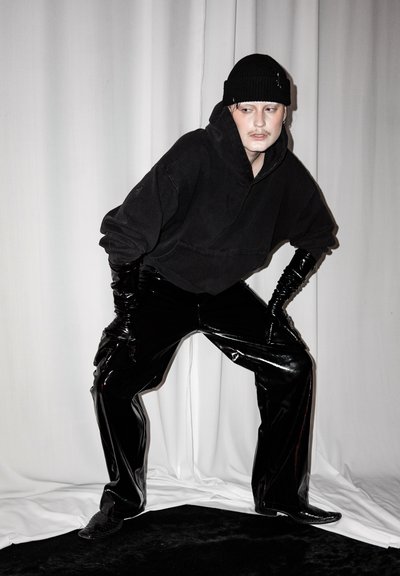 Persona que lleva un gorro negro, una sudadera negra extragrande, pantalones negros de vinilo brillantes y guantes negros largos, posando con las manos en las rodillas sobre un fondo blanco.