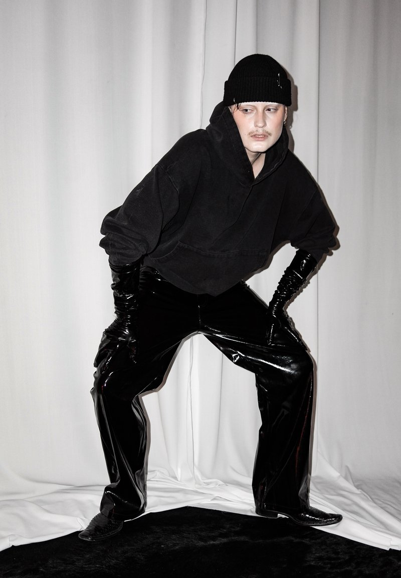 Άτομο που φοράει μαύρο σκουφί, oversized μαύρο φούτερ, γυαλιστερό μαύρο βινυλικό παντελόνι και μακριά μαύρα γάντια, ποζάρει με τα χέρια στα γόνατα σε λευκό φόντο.