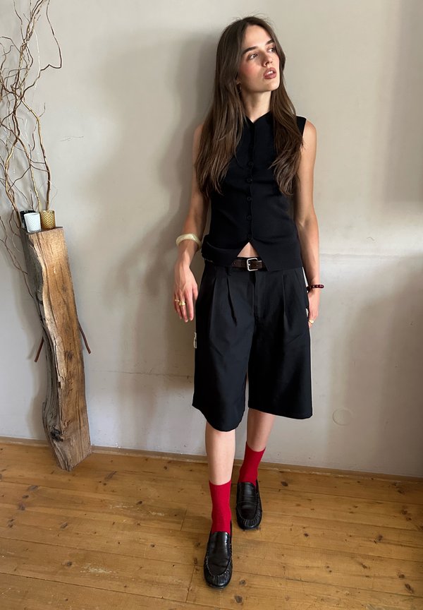 Junge Frau mit langen Haaren, die ein schwarzes ärmelloses Oberteil, schwarze lange Shorts, rote Socken und schwarze Schuhe trägt und drinnen an einer Wand steht.