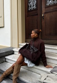 Kvinne i brun rullehalsgenser og lysebrune støvler som sitter på steintrappen og holder en stor mørkebrun veske ved en tre dør.