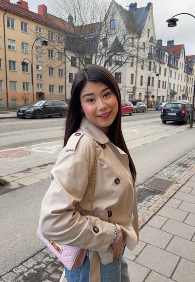 Joven mujer con cabello largo y oscuro, vistiendo un abrigo beige y jeans azules, sonriendo en una acera de la ciudad con coches estacionados y edificios al fondo.