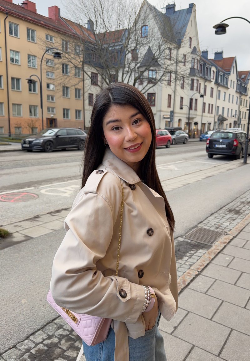 Młoda kobieta o długich ciemnych włosach ubrana w beżowy płaszcz i niebieskie dżinsy, uśmiecha się na miejskim chodniku, w tle zaparkowane samochody i budynki.