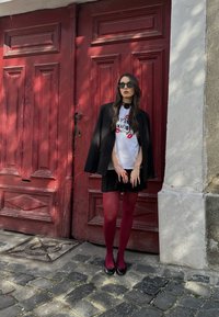 Czarna marynarka noszona na białej koszulce z odciskami warg, zestawiona z czarną mini spódnicą i burgundowymi rajstopami. Stoi przy czerwonych drzwiach.