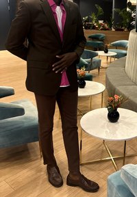 Ruskea räätälöity bleiseri kirkkaan pinkin paidan päällä, yhdistettynä ruskeisiin istuviin housuihin ja tummiin loafer-kenkiin. Sisätilassa, jossa on modernia huonekalua.