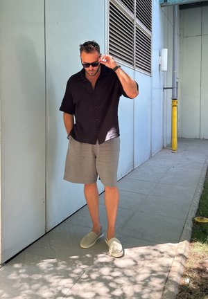 Hombre con una camisa negra de manga corta y botones, pantalones cortos grises y alpargatas claras, caminando por un sendero pavimentado junto a una pared azul.