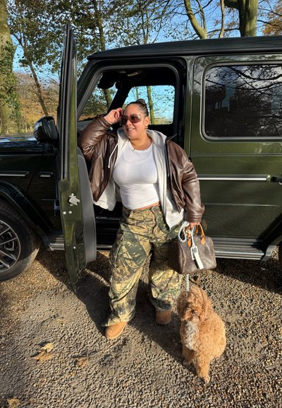 Chaqueta de cuero marrón sobre un top corto blanco, pantalones de camuflaje, sosteniendo un bolso marrón, de pie junto a un SUV negro con un perro marrón.