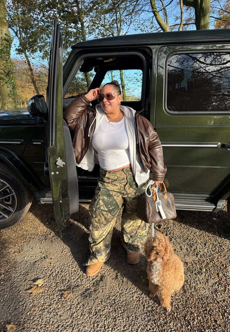 Veste en cuir marron sur un crop top blanc, pantalon camouflage, tenant un sac à main marron, debout à côté d'un SUV noir avec un chien marron.
