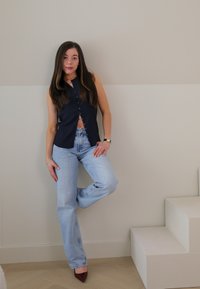 Mornarsko brezrokavno zgornje oblačilo z gumbi, svetlo modri ravni kavbojke in pointed burgundski čeveljci, postavljeni ob nevtralno steno in minimalističnih stopnicah.