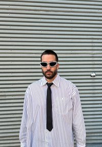Άσπρη και μαύρη ριγέ μπλούζα με μακριά μανίκια, μαύρη γραβάτα και άσπρα γυαλιά ηλίου, στήνεται μπροστά από φόντο γκρίζου σιδερένιου μετάλλου.