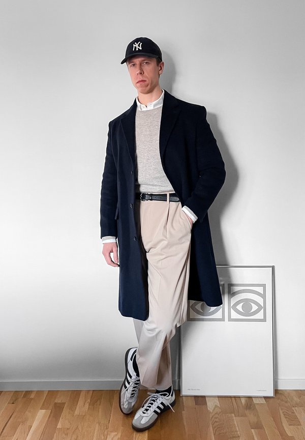 Man draagt een marineblauwe jas, grijze trui, beige broek, zwarte riem, sneakers en zwarte pet en staat binnen tegen een witte muur met ingelijste oogkunst.