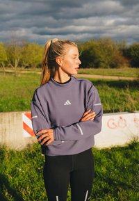 Jonge vrouw met blond haar in een paardenstaart, gekleed in een paarse sporttop met lange mouwen en zwarte legging, staande in de buitenlucht met haar armen gekruist.