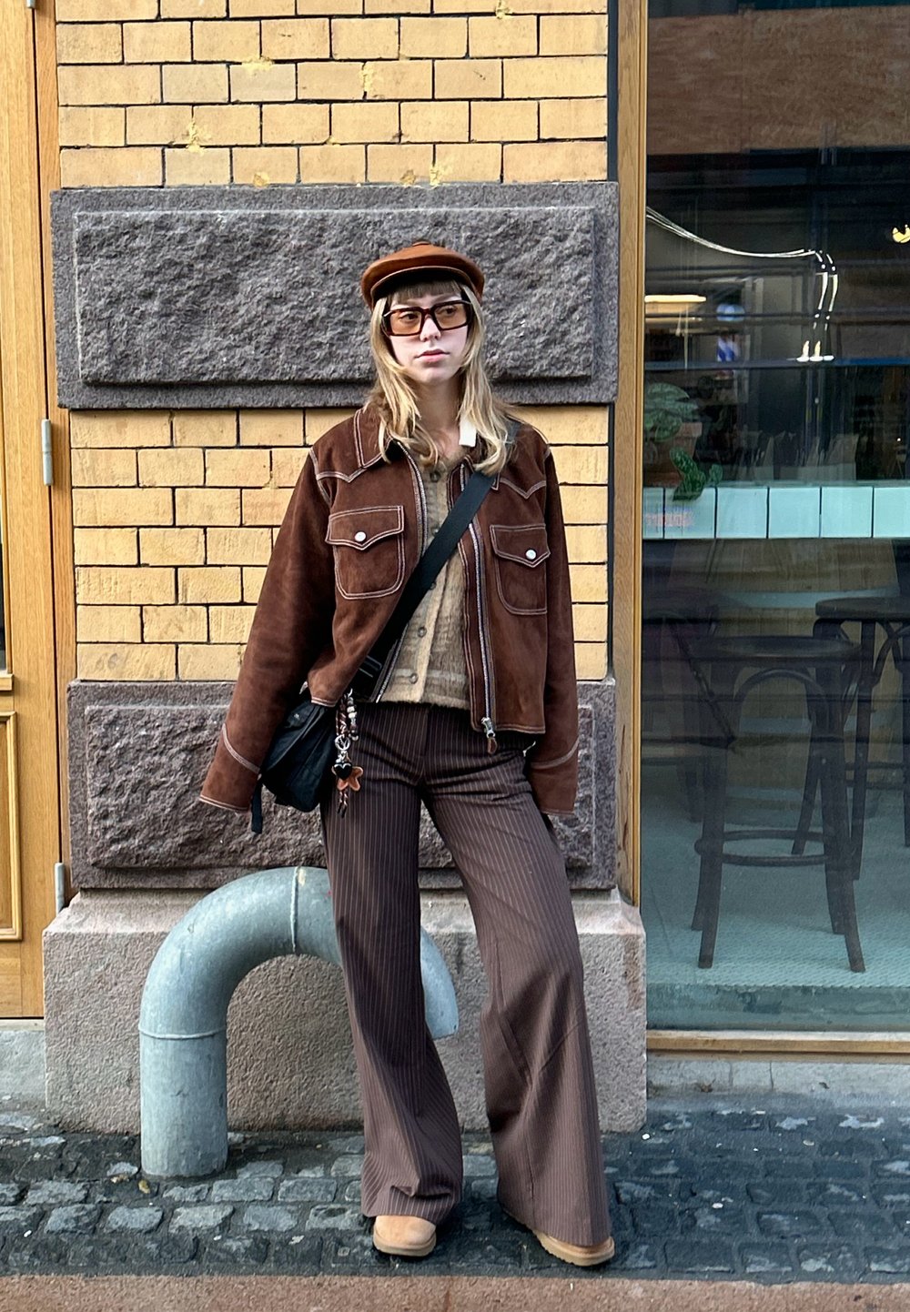 Hnědá semišová bunda s bílým prošitím, béžový svetr, hnědé kalhoty s širokými nohavicemi a proužky, hnědé kozačky a hnědá čepice. Černá kabelka přes rameno.