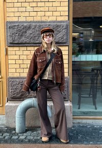 Hnědá semišová bunda s bílým prošitím, béžový svetr, hnědé kalhoty s širokými nohavicemi a proužky, hnědé kozačky a hnědá čepice. Černá kabelka přes rameno.