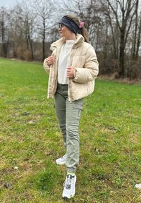 Casaco puffer bege por cima de um suéter de malha branco, calças cargo verdes e ténis brancos. O cenário é um campo gramado com árvores despidas.