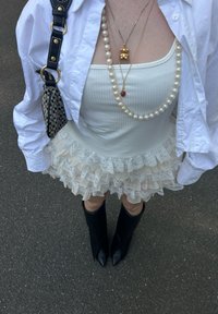 Top de alças branco texturizado por baixo de uma camisa branca abotoada, saia bege com camadas e volantes, colar longo de pérolas, charme dourado, botas pretas até ao joelho.