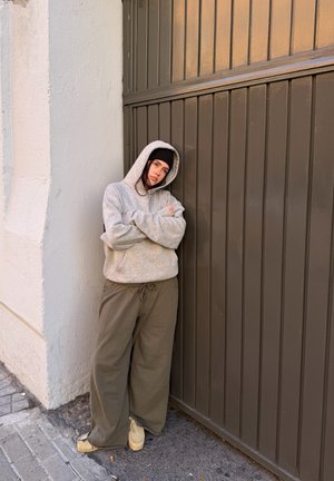 Persona joven con sudadera con capucha gris, gorro negro y pantalones ancho de color oliva se apoya contra una puerta marrón panelada con los brazos cruzados en una acera urbana.