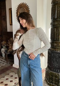 Blouse à manches longues crème clair avec une texture froncée, assortie à un jean en denim large à taille haute. Sol à motifs aux teintes neutres en arrière-plan.