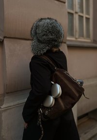 Grå pelslue, svart kåpe, brun semsket skinnveske med sølvfargede hodetelefoner festet. Teksturene er myke og kontrasterende. Bakgrunnen har en lys vegg.