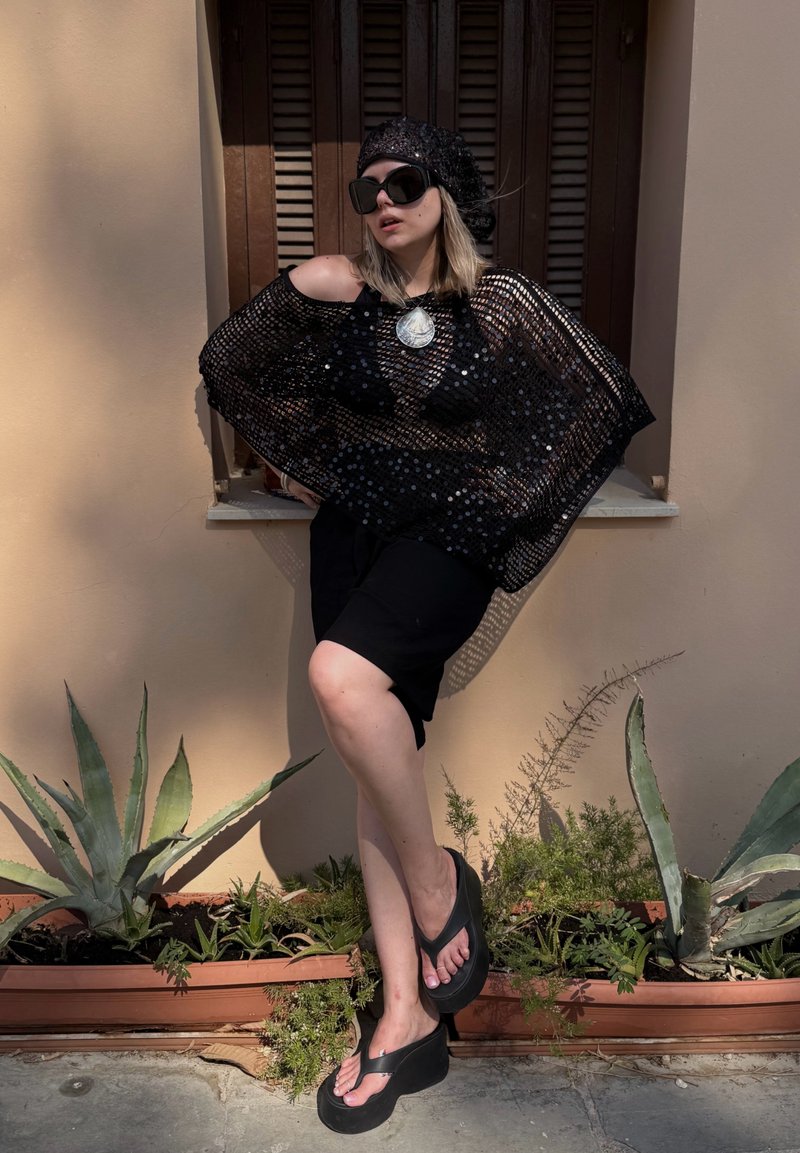 Femme portant un haut en résille noir, un short noir, des tongs noires, un béret à sequins et de grandes lunettes de soleil, appuyée contre un mur beige avec des plantes en dessous.