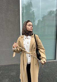 Beige trenčkot s opaskou v pase přes bílé tričko. Leopardí šátek kolem hlavy. Černá kabelka přes rameno, stojící proti texturované zdi.