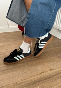 Sort og hvid sneaker med ruskind og lædermaterialer, der har tre hvide striber, en gummisål og et guldlåg på tungen.