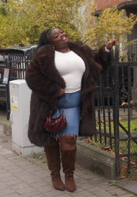 Femme confiante portant un manteau en fourrure marron, un haut blanc, une jupe en denim et des bottes marron, debout sur le trottoir, tenant un sac rouge et appuyée contre une clôture en fer noir.