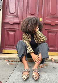 Luipaardprint jasje, zwarte top, grijze wijde broek, puntige luipaardprint schoenen met gespen, zittend tegen een kastanjebruine deur.