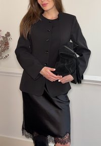 Γυναίκα που φοράει μαύρο σακάκι με κουμπιά και σατέν φούστα με δαντελένιο τελείωμα, κρατώντας μια μαύρη τσάντα μπροστά σε λευκό τοίχο με ξεραμένα λουλούδια κοντά.