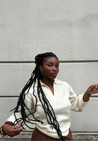 Maglione a maglia testurizzata color crema chiaro con scollatura a V e maniche 3/4, abbinato a pantaloni marroni a vita alta e lunghe trecce nere.