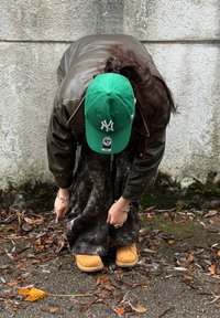 Hnedá kožená bunda, zelená čiapka s bielym logom, voľné čierne nohavice s vzorom, svetlohnedé topánky, ruky držia nohavice, v pozadí listie.