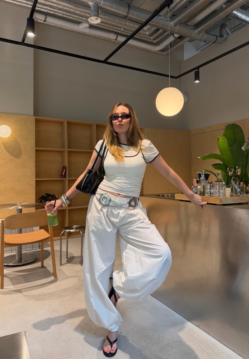 Femme en pantalons larges blancs et t-shirt, portant des lunettes de soleil, tenant une boisson, appuyée sur un comptoir dans un café moderne au décor minimaliste.
