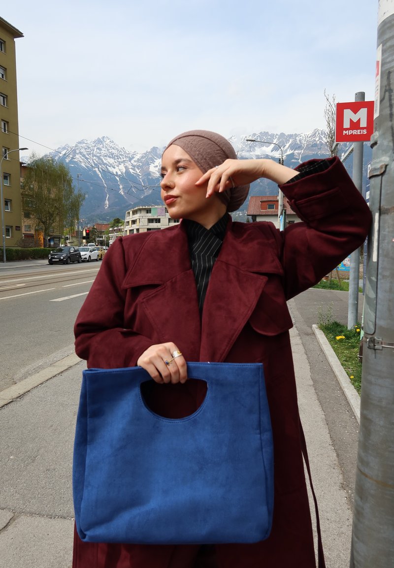 Vrouw in een kastanjebruine jas en hoofddoek houdt een grote blauwe tas vast terwijl ze op straat staat met bergen en gebouwen op de achtergrond.