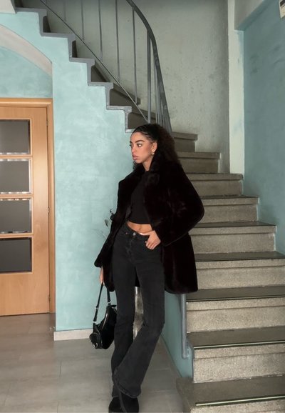 Abrigo de piel sintética negra, top negro corto y jeans negros de campana. Escalera curva al fondo, pared azul clara, puerta de madera.