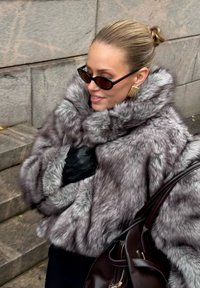 Szara kurtka z sztucznego futra z wysokim kołnierzem, czarne skórzane rękawice i ciemne okulary przeciwsłoneczne. Kurtka ma grubą, miękką teksturę i luźny krój.