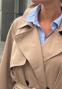 Bež trench coat kahekordse disainiga, suure kraega ja mustade nuppudega. Kanda helesinise, triibulise kraejoonega särgi kohal.