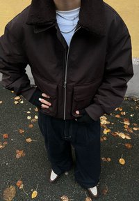 Tamno smeđa bomber jakna s teksturiranim smeđim ovratnikom od flisa, zatvaračem i dva prednja džepa, uparena s opuštenim tamnoplavim trapericama i bijelim cipelama.