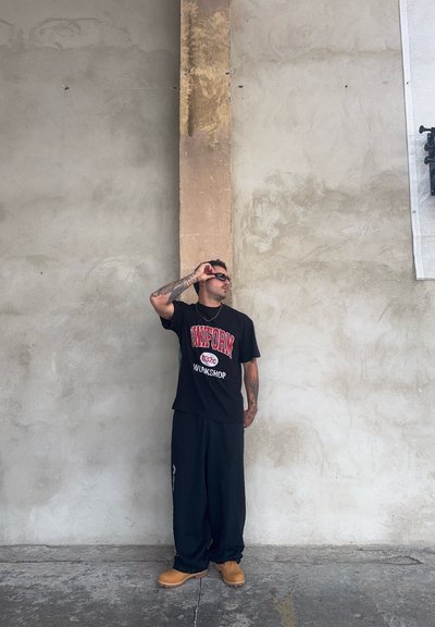 Camiseta negra con texto gráfico rojo, pantalones negros holgados y zuecos marrón claro. Tatuajes visibles en los brazos. Fondo neutro con pared texturizada.