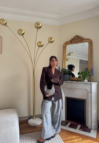 Blazer brun avec sac noir et gris ; pantalon large gris clair ; lampe sur pied dorée ; cheminée en marbre avec miroir orné et plantes en pot.