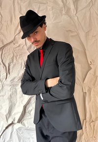 Svart kostym med röd slips, bärs över en skjorta med krage. Personen har på sig en svart fedora-hatt och står med armarna i kors mot en texturerad bakgrund.