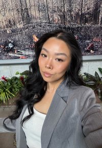 Nuori nainen, jolla on pitkät mustat hiukset, pukeutuneena harmaaseen bleiseriin ja valkoiseen puseroon, poseeraa sisätiloissa kasvien ja tekstuuriseinän edessä.