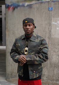 Camo-jack met een zwart shirt met een schedelontwerp, gecombineerd met rode broek. Staand tegen een betonmuur.