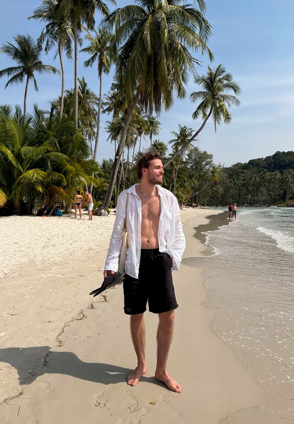 Man in open white shirt and black shorts stands barefoot on sandy beach holding sandals, with palm trees and ocean waves in the background.