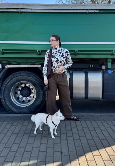 Persona vistiendo una chaqueta de estampado de vaca en blanco y negro y pantalones anchos marrones se encuentra junto a un perro blanco con correa, cerca de una camioneta verde.