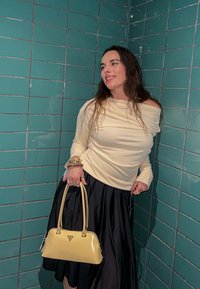 Kreemjas pika varrukaga topp langeb ühe õla peale, musta volditud seelikuga. K�äes kollane käekott kahe käepidemega. Taustal sinakashall ruuduline plaat.